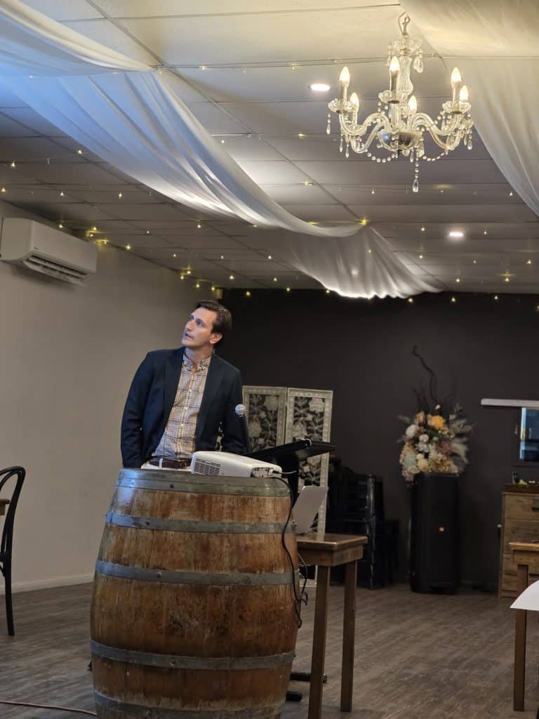 Man speaking at event with chandelier and draped ceiling decor.